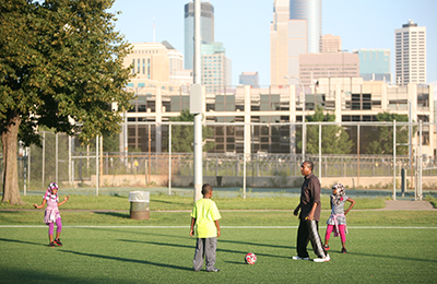 Adult and three kids playing soccer on a field with downtown Minneapolis in the background